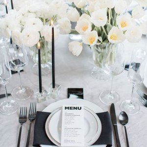 Elegant table setting with white flowers, black candles, and a menu on top of a plate, surrounded by cutlery, glassware, and folded black napkins.