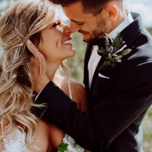 A bride and groom are standing close, smiling at each other. The groom is wearing a black suit, and the bride is in a white dress. They appear to be outdoors.