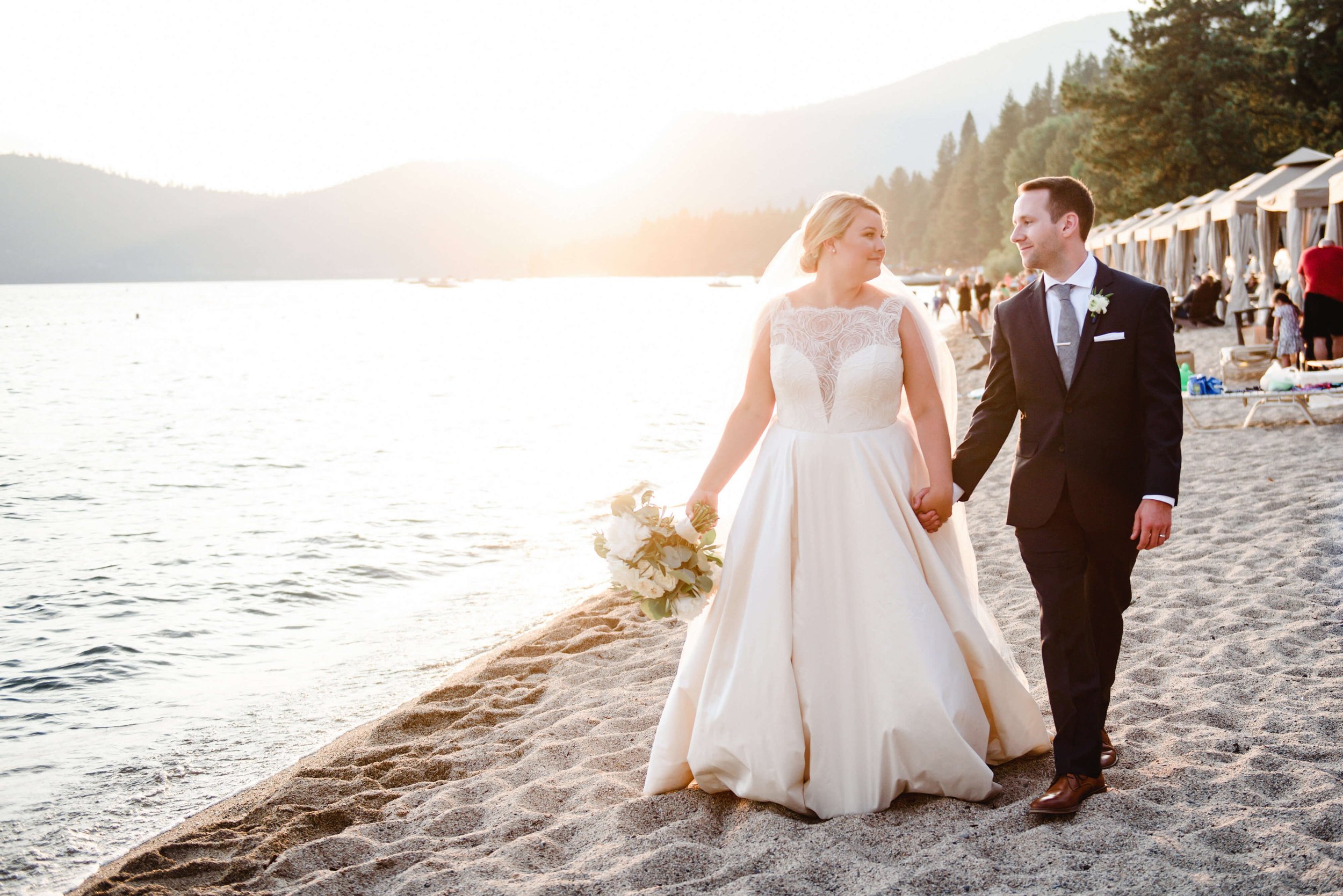 A bride and groom walk hand-in-hand along a sandy beach at sunset, with the bride in a white dress holding a bouquet and the groom in a dark suit. Trees and mountains are visible in the background.