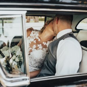 A bride and groom share a kiss inside a car, with the bride wearing a white lace dress and holding a bouquet.