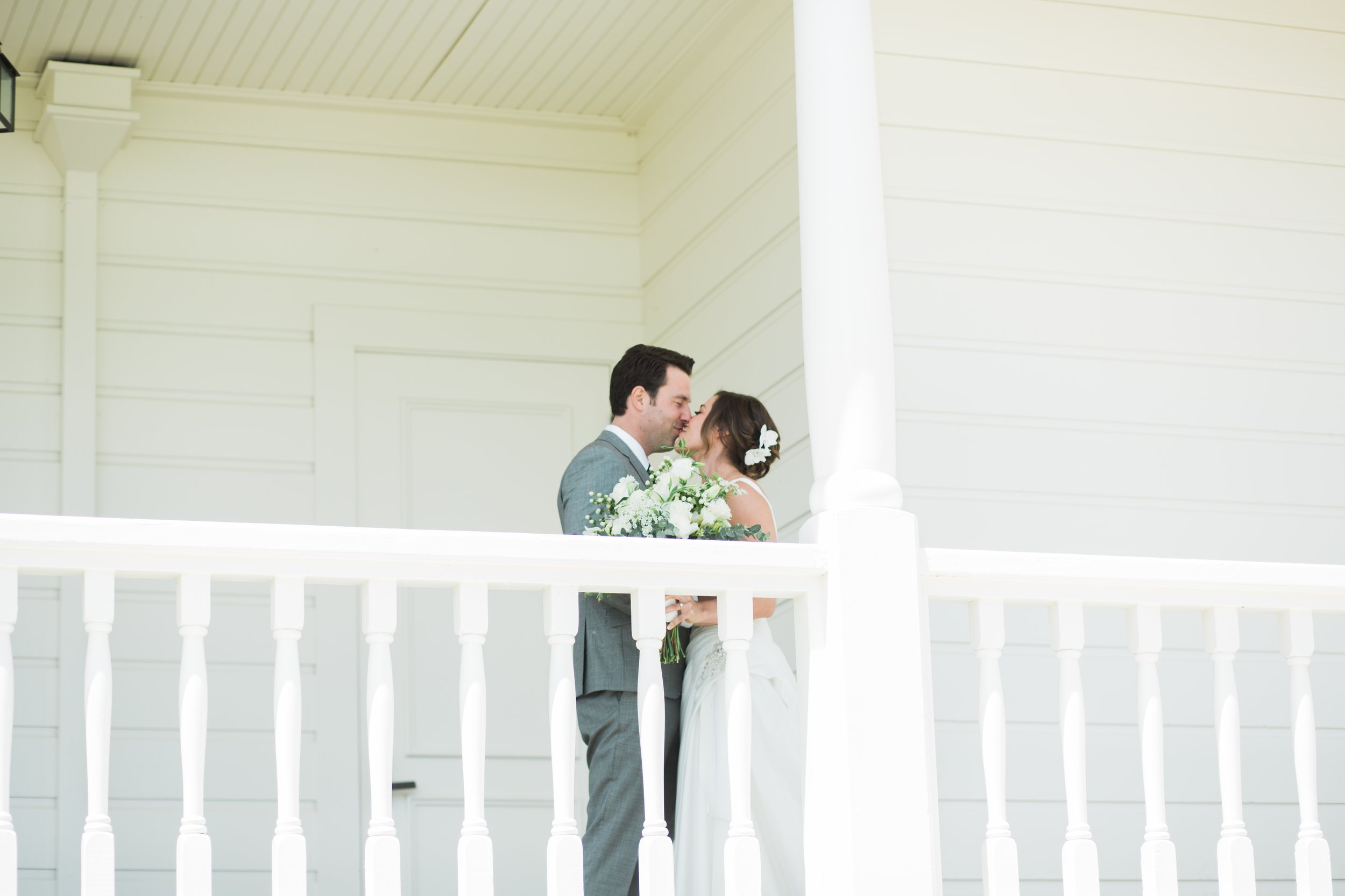 A couple in wedding attire shares a kiss on a white porch, holding a bouquet of flowers.