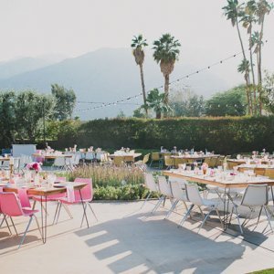 Outdoor dining setup with multiple tables and chairs, decorated with flowers and set against a backdrop of palm trees and mountains. String lights hang overhead.