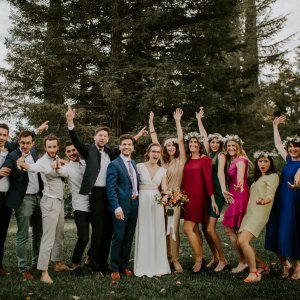 A group of people, including a bride in a white dress and a groom in a blue suit, pose enthusiastically in front of trees. Some are wearing flower crowns.