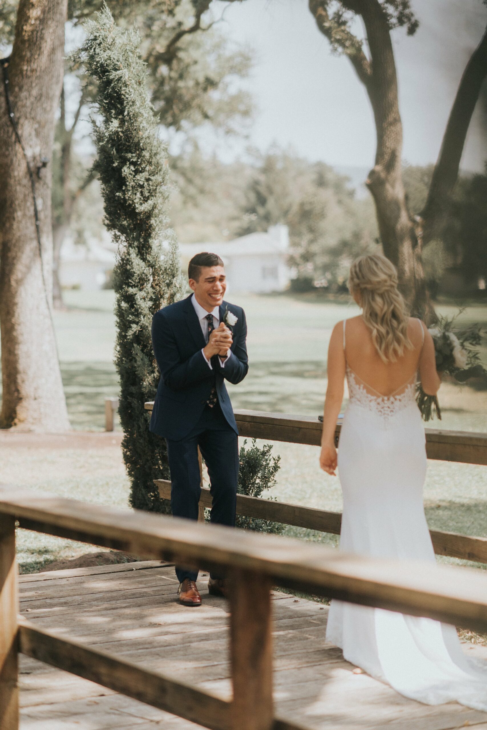 A bride and groom stand on a wooden platform outdoors. The groom, in a navy suit, holds a microphone and appears to be speaking, while the bride in a white dress faces him holding a bouquet.