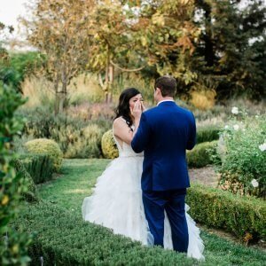 A bride in a white gown and a groom in a blue suit stand facing each other in a lush garden setting during their wedding ceremony.