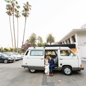 A couple dressed in wedding attire sits on the step of a white van in a parking lot with palm trees and several cars in the background. They are leaning in to kiss.