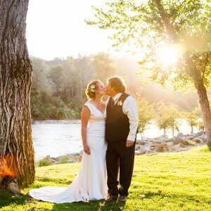 A couple dressed in wedding attire shares a kiss outdoors by a river, surrounded by trees and illuminated by sunlight.