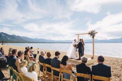 A couple stands under a wooden arch on a sandy beach during their wedding ceremony, with seated guests watching and a mountainous backdrop visible.