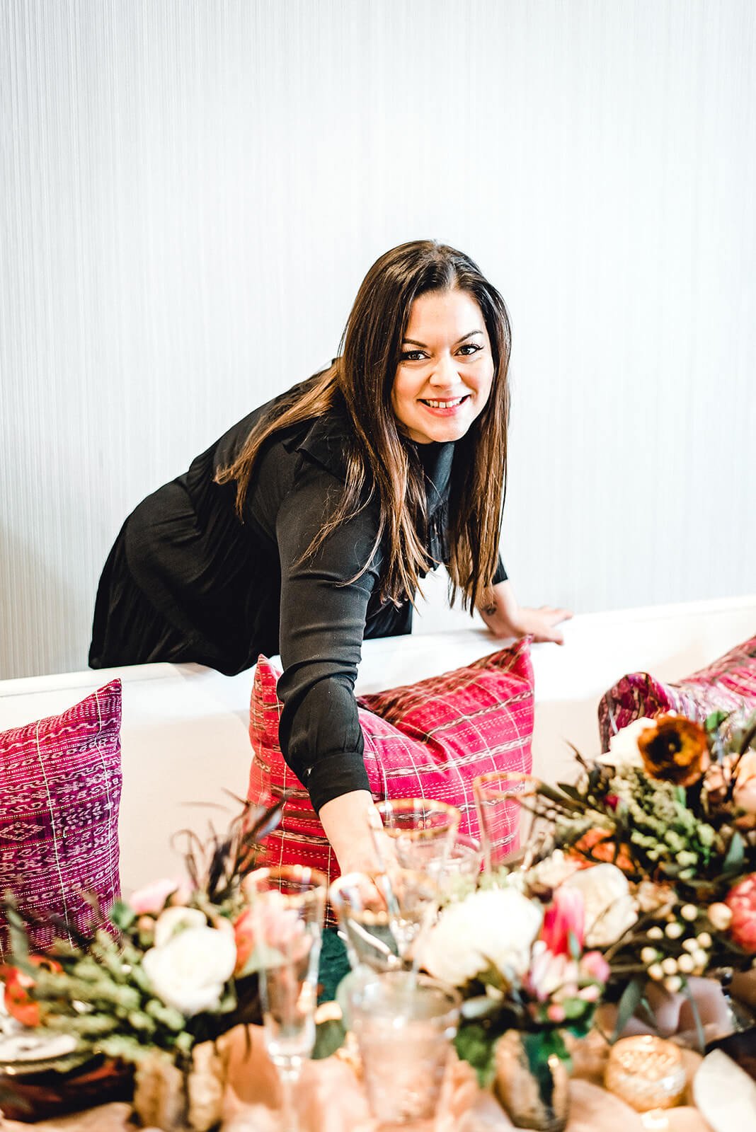 Woman in a black blouse setting a table adorned with flowers, glassware, and colorful pillows.