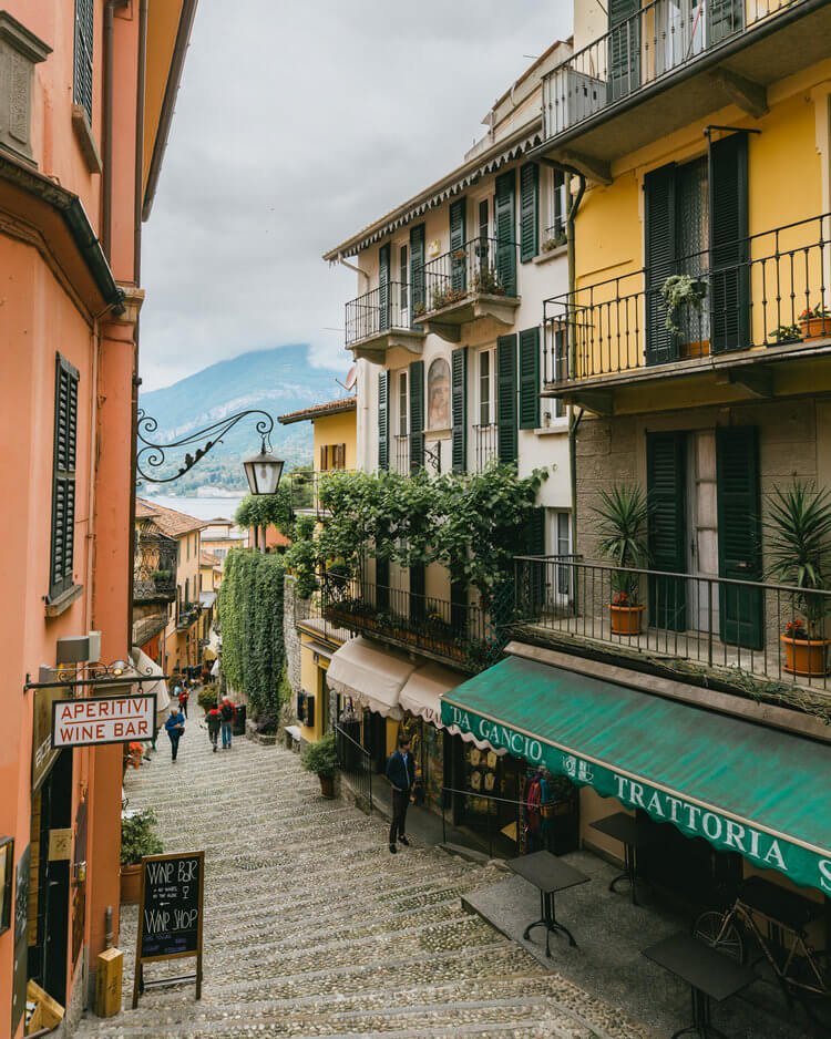 A narrow, cobblestone street lined with colorful buildings, shutters, balconies, and signs for a wine bar and trattoria, with mountains visible in the background.