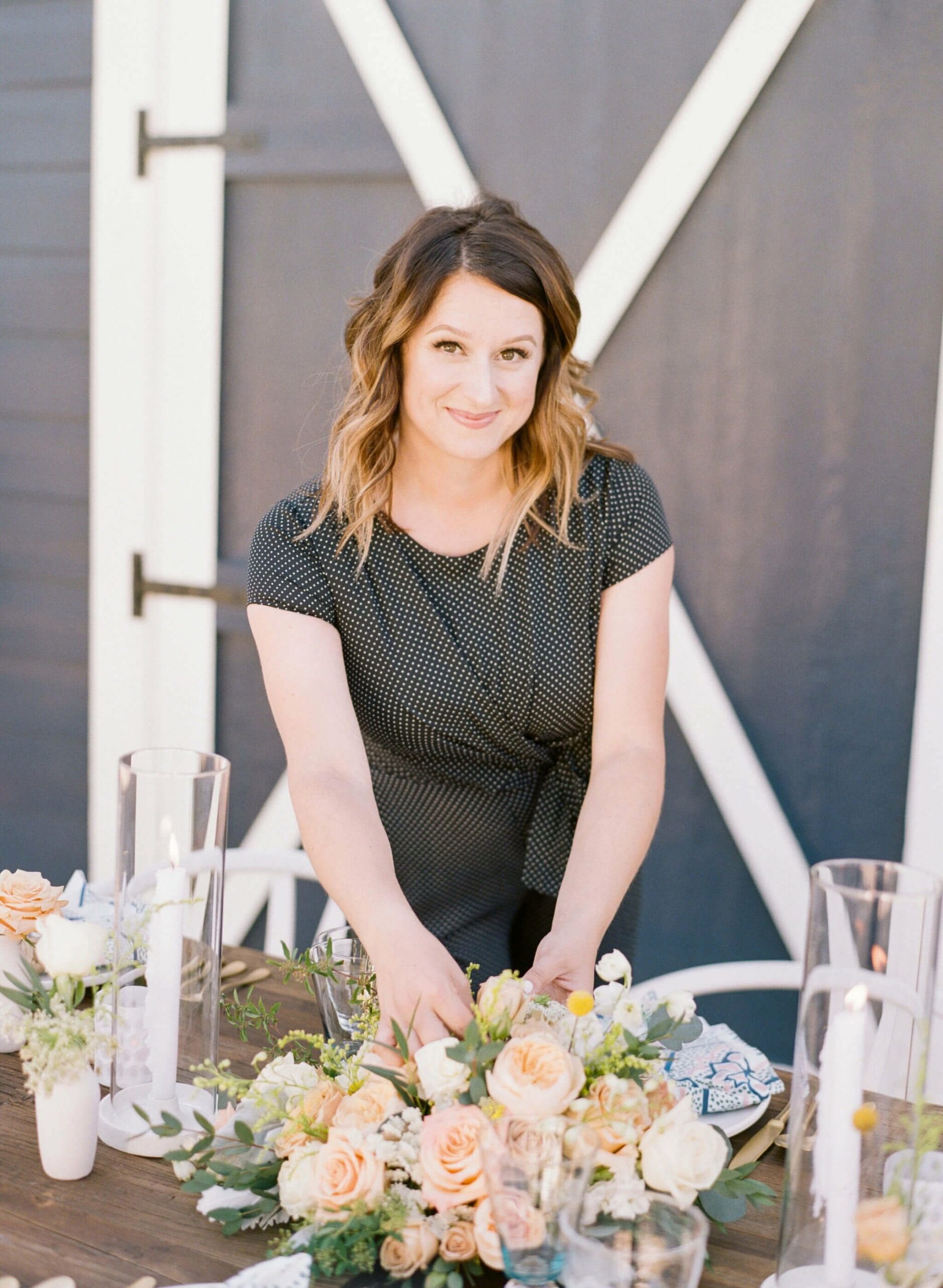 A person arranging flowers on a table set with candles and floral arrangements in front of a barn door.