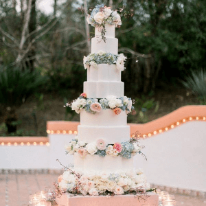 A five-tier wedding cake decorated with pastel flowers sits outdoors with trees and lit decorations in the background.