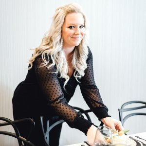 A woman with long, wavy blond hair wearing a black, sheer polka-dotted blouse is setting up a table with chairs around it.