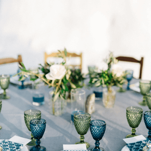 A table is set with blue and green glass goblets, floral centerpieces, and place cards on a gray tablecloth.