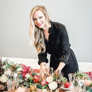 A smiling person with long blonde hair, wearing a black dress, arranging floral decorations on a table.