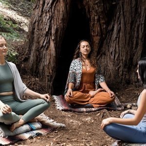 Three women sit cross-legged on blankets placed on the ground in front of a large tree, engaged in a meditation session in a forest setting.