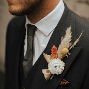 Close-up of a man wearing a dark suit, white shirt, and patterned tie. His lapel is adorned with a boutonniere featuring various flowers and foliage.