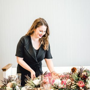 A person with long hair in a black outfit arranges floral decorations on a table.