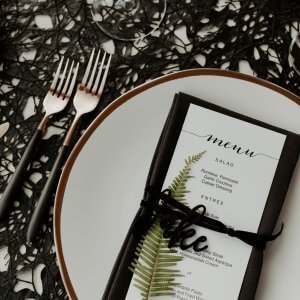 A table setting with a white plate, black utensils, and a menu wrapped in black ribbon featuring a fern design, displayed on a decorative black tablecloth.