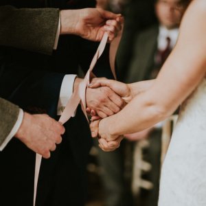 A close-up of a couple's hands being tied together with pink ribbons during a wedding ceremony.