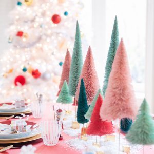 A dining table is decorated with miniature pastel Christmas trees, plates, and festive ornaments. In the background, a brightly lit Christmas tree is adorned with colorful baubles.