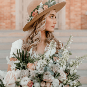 A woman with long, wavy blonde hair, wearing a hat adorned with flowers, holds a large bouquet of assorted flowers, standing in front of steps and a brick wall.