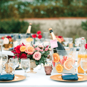 Elegant table setting with gold plates, blue napkins, and floral centerpieces. Champagne bottles in ice buckets with citrus slices, glasses, and bottles of juice on a white tablecloth.