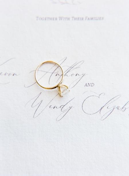 A gold ring with a diamond is placed on a white paper with faint, cursive text in the background.