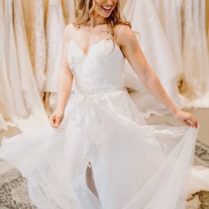 A woman in a white wedding dress smiles and holds the sides of her dress in a bridal store.