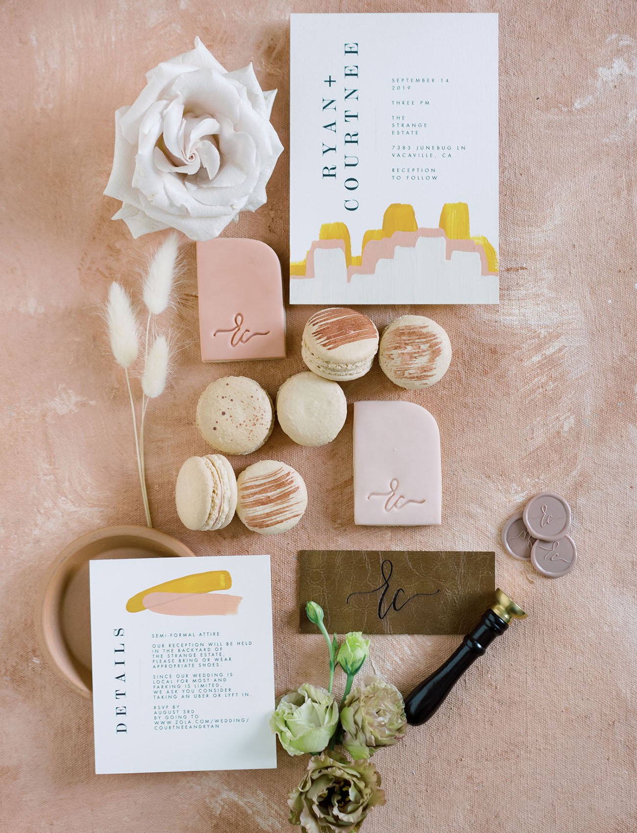 Assorted wedding stationery and desserts, including invitations, macarons, wax seals, and floral decor, arranged on a peach background.