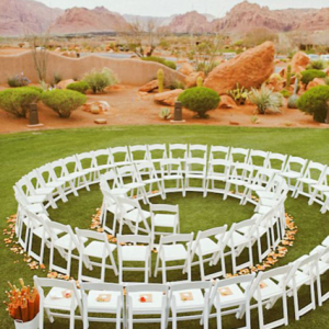 Rows of white folding chairs arranged in concentric circles on green grass, with a backdrop of reddish desert landscape and rocky formations.