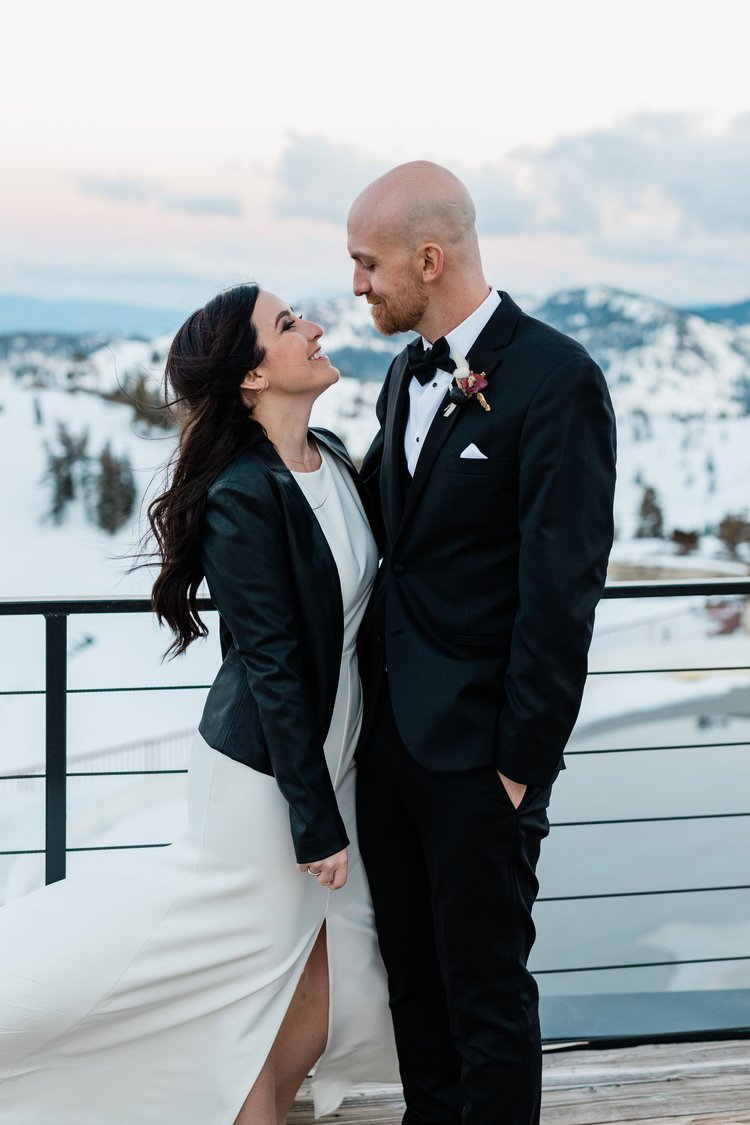 A couple dressed in formal attire, the woman in a white dress and black jacket, and the man in a black suit, stand closely together on a balcony overlooking a snowy landscape.