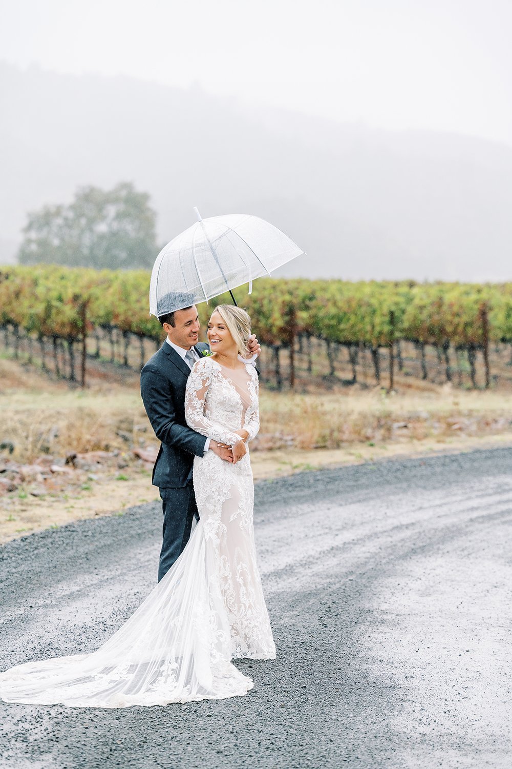 A bride and groom stand on a wet road in front of vineyards, sharing a transparent umbrella on a rainy day, both smiling and dressed in wedding attire.