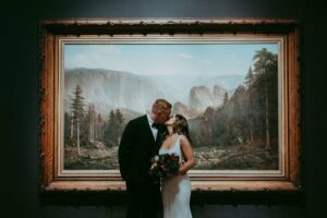 A bride and groom kiss in front of a large framed landscape painting, with trees and mountains in the background.