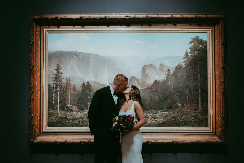 A bride and groom kiss in front of a large framed landscape painting, with trees and mountains in the background.