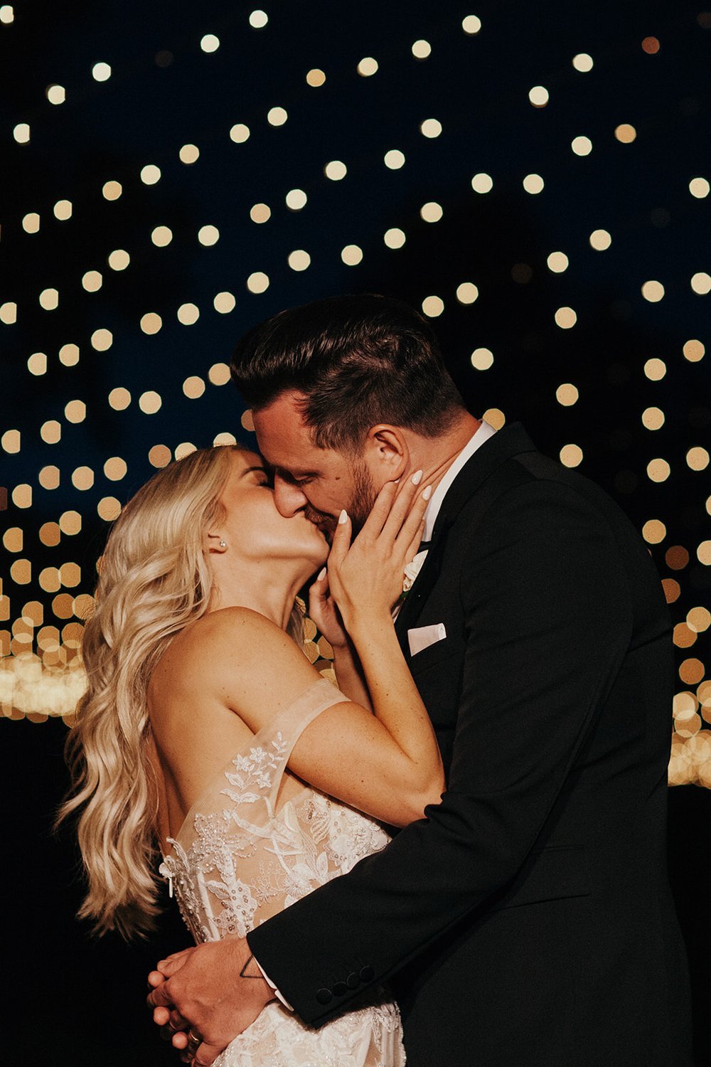 A couple in wedding attire shares a kiss under a night sky filled with blurred lights.