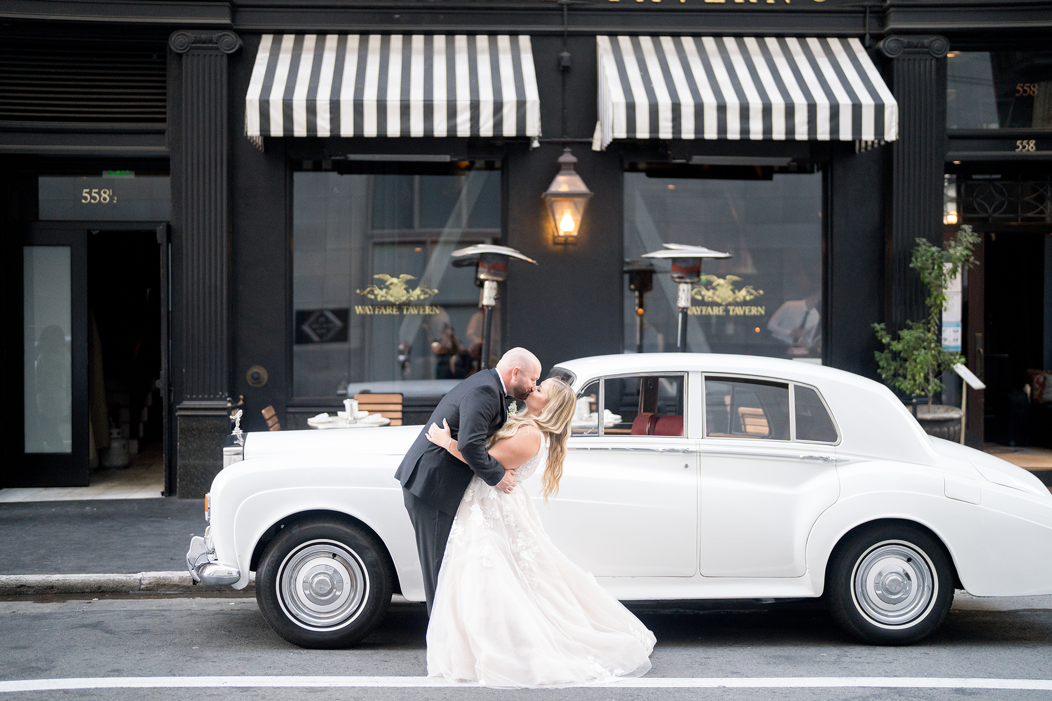 A couple in wedding attire share a kiss in front of a vintage white car parked outside a building with striped awnings.