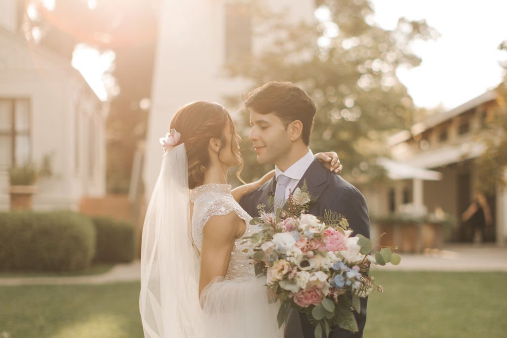 A bride and groom in wedding attire share an intimate moment outdoors, with the bride holding a colorful bouquet.