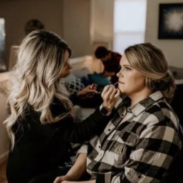 A woman in a checkered shirt sits while another woman with long blonde hair applies makeup to her face in a well-lit room.