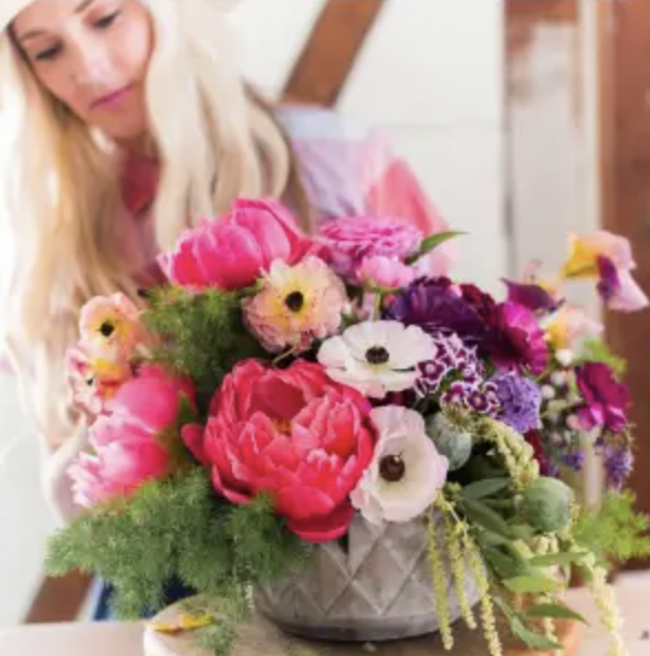 A person with long, blonde hair arranges a bright floral bouquet with pink, white, and purple flowers in a textured vase.