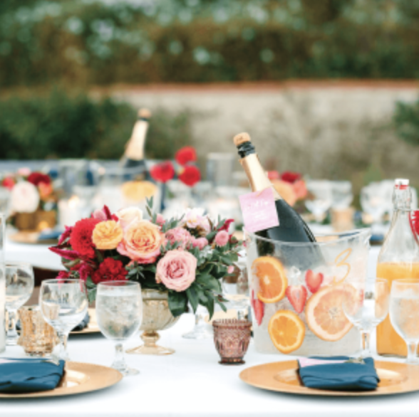 A table set for an outdoor event featuring floral centerpieces, champagne bottles in ice buckets, glassware, plates with blue napkins, and a juice bottle.