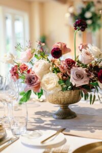 A floral arrangement in a gold vase features a mix of pink, white, and dark red roses on a banquet table set with glassware and plates.