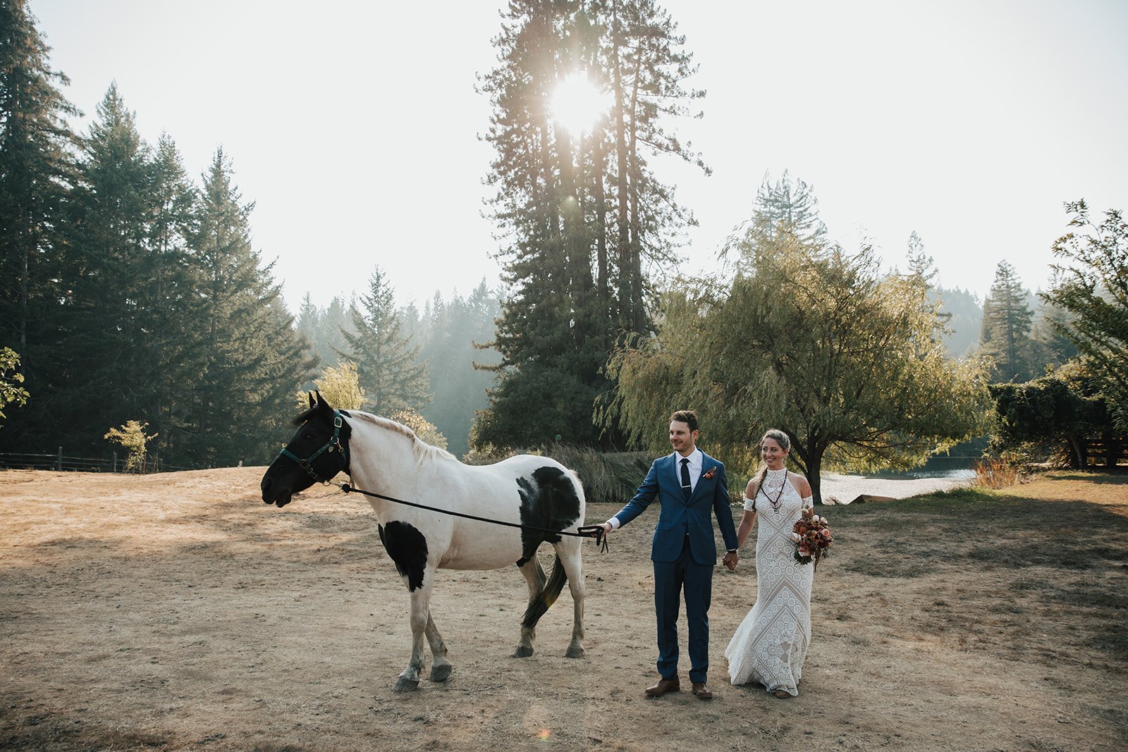 A couple in wedding attire holds hands while walking a black and white horse in a sunlit, wooded area.