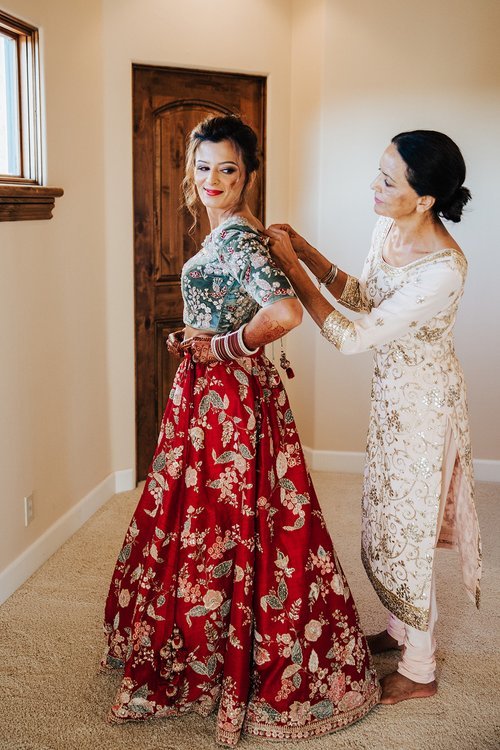 A woman stands in a decorated red dress while another woman helps tie the back of her blouse in a brightly lit room.