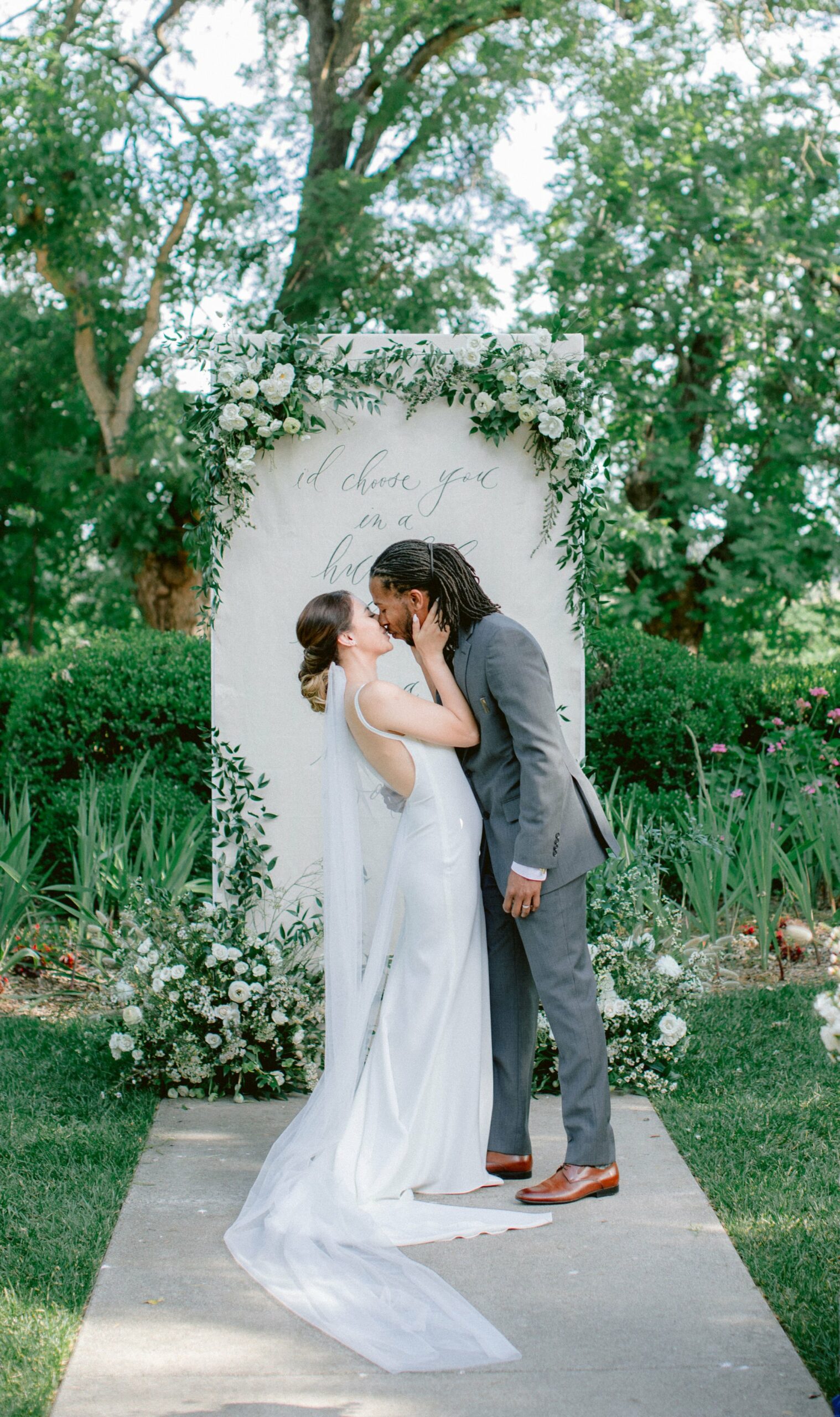 A bride and groom kiss in front of a floral archway outdoors, with text on a backdrop behind them. Both are dressed in formal wedding attire.