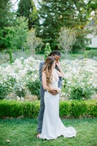 A couple stands closely in a garden, the woman in a white dress and the man in a gray suit, surrounded by greenery and white flowers.