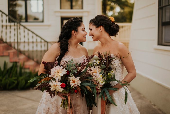 Two women in sleeveless dresses hold bouquets and face each other, nose to nose, outside a building with a staircase and greenery in the background.