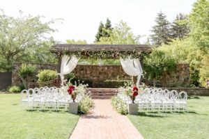 An outdoor wedding altar decorated with draped white fabric and flowers is set up in a garden with white chairs arranged in rows on a grassy area and a brick walkway leading to the altar.