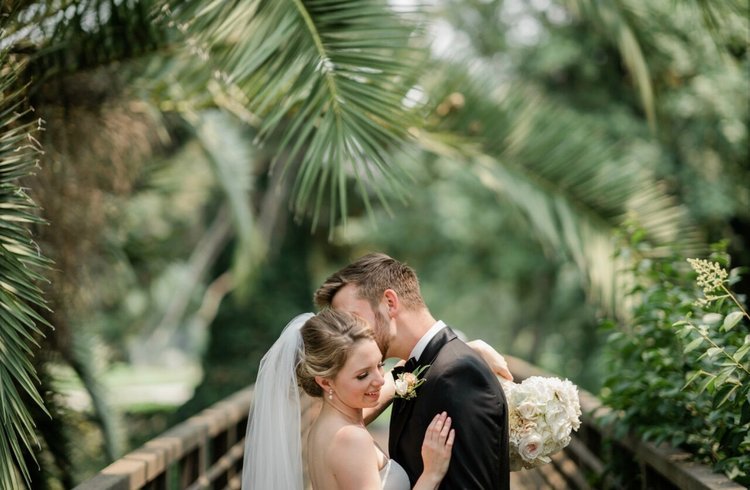 A couple dressed in wedding attire embraces on a wooden bridge surrounded by lush greenery and palm leaves. The woman holds a bouquet, and both appear content.