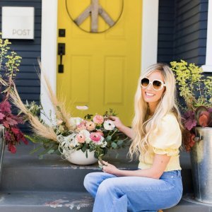 Woman with long blonde hair wearing sunglasses sits on the steps of a house with a bright yellow door, arranging flowers in a vase.